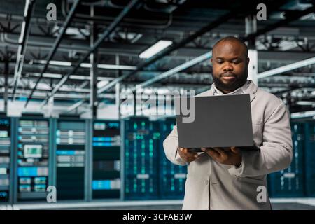 Technician in server hub managing infrastructure systems, checking recovery plan on laptop. Data center employee monitoring performance using tools, making sure rigs are functioning optimally Stock Photo