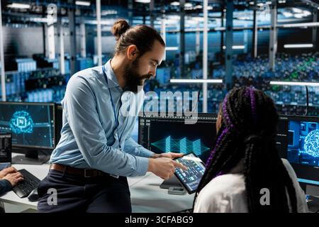 Team leader guides intern in data center to troubleshoot server connectivity issue using AI. Mentor teaches trainee during internship to audit systems for vulnerabilities using artificial intelligence Stock Photo