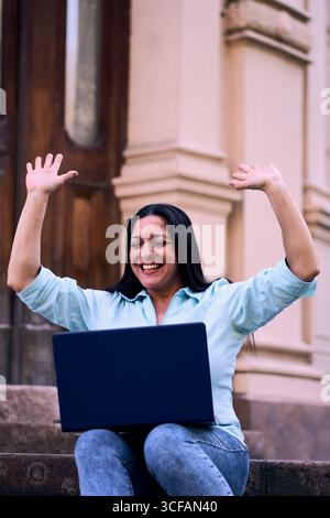 Beautiful mature Latina woman working on her laptop outdoors at home, smiling, energetic, and very happy with her arms raised. Businesswoman Stock Photo