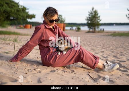 Cat watching woman owner relaxing on sandy beach on seaside walk. Shore stroll with pet feline Stock Photo