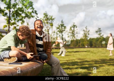 Caucasian boy and Black boy sitting outdoors using digital tablets, Black boy laughing while Caucasian boy looking down at device, girls playing in background Stock Photo