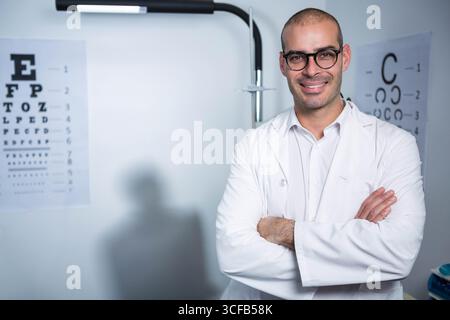 Male optometrist in lab coat standing in exam room beside Snellen chart, C-shaped chart, copy space Stock Photo