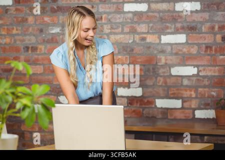 Woman in her twenties leaning over silver laptop on wooden desk featuring potted plants in office Stock Photo