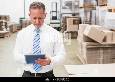 Mature man standing in warehouse wearing tie holding tablet by forklift and pallets checking stock Stock Photo