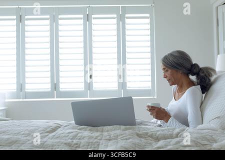 Senior woman sitting on bed near window shutters holding white mug while using laptop, copy space Stock Photo