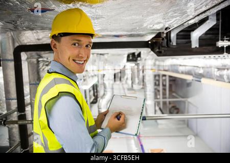 Male engineer wearing helmet, vest checking ducts in hall holding clipboard, pen, copy space Stock Photo