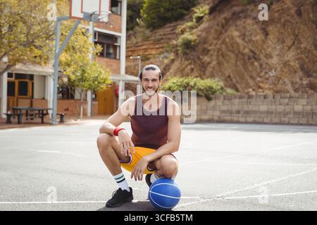 Adult man in sportswear kneeling at outdoor basketball court gripping ball near metal hoop Stock Photo