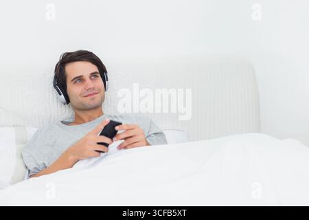 Man reclining on bed in bright bedroom, holding smartphone and wearing headphones, copy space Stock Photo