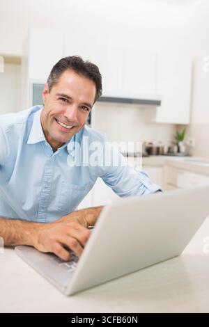 Middle-aged man in casual shirt leaning over kitchen counter using silver laptop next to spice jars Stock Photo