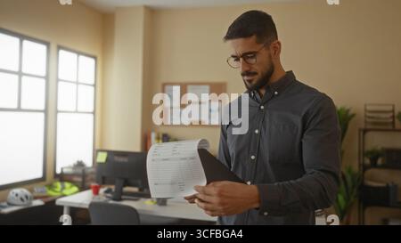 Young man in an office setting reviews documents attentively, surrounded by office furniture, creating a focused workplace atmosphere. Stock Photo