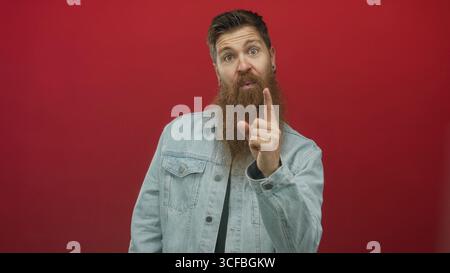Man with long beard wearing denim jacket over black shirt waves hand in red studio looking up; happiness. Stock Photo