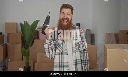 Bearded man holds cordless drill and smiles inside building stacked with cardboard moving boxes in new home; excitement achievement. Stock Photo
