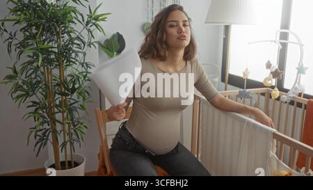 Pregnant woman sitting in a light-filled nursery cooling herself with a paper fan beside a crib surrounded by baby decor and plants, looking thoughtfu Stock Photo