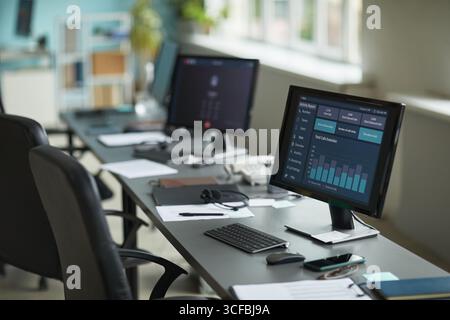 Row of empty office desks with computer monitors displaying data analytics dashboard, keyboards and mice arranged on workstations, modern workspace illuminated by natural light from large windows Stock Photo