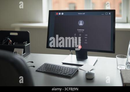 Modern office desk featuring computer monitor displaying voice call interface with customer profile, wireless keyboard and mouse placed on table, window in background Stock Photo