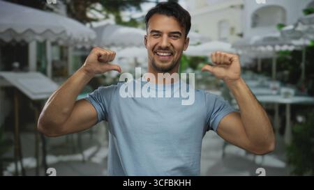 Young hispanic man on terrace pointing to himself with thumbs smiling at outdoor restaurant setting under umbrellas. Stock Photo