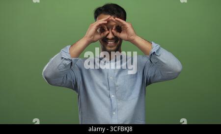 Smiling young hispanic man in casual blue shirt holding hands like binoculars over eyes against isolated green background. Stock Photo