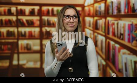 Woman holding smartphone in library with blonde hair and eyeglasses, showcasing a reflective mood under warm lighting among rows of bookshelves indoor Stock Photo