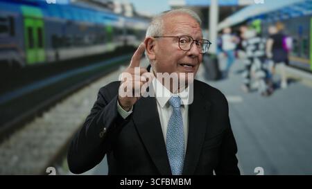 Senior man in suit at outdoor train station pointing upwards with blurred train and commuters in background. Stock Photo