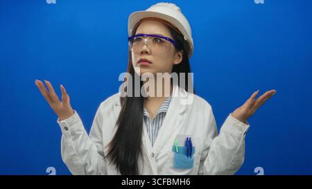 Young chinese woman in hardhat and lab coat raises hands in question against isolated blue background, illustrating confusion or inquiry. Stock Photo