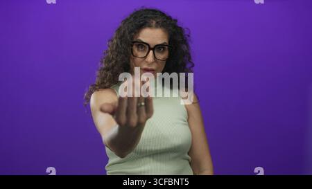 Young woman wearing glasses and green top beckons hand forward in purple studio; friendly invitation. Stock Photo