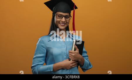 Woman graduate smiling, wearing graduation cap, holding book against vibrant orange background, embodying academic success and happiness. Stock Photo