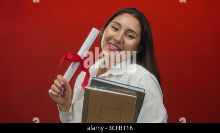 Young hispanic woman holds diploma with red ribbon and stack of books in studio; proud achievement. Stock Photo