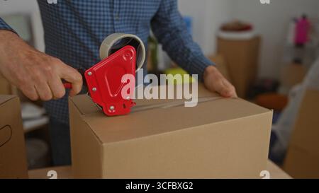 Man sealing cardboard box in home interior using red tape dispenser, representing moving or relocation process with packaging materials in living room Stock Photo