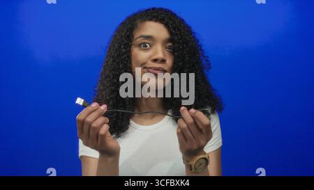 African american woman holding a broken usb cable with hands in a vibrant blue studio; frustration connection failure. Stock Photo