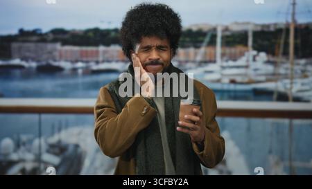 Man stands outdoors by the seaside holding coffee near boats, expressing tooth pain while touching jaw against a backdrop of a port and calm sea. Stock Photo