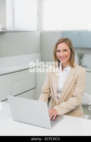 Senior woman wearing beige blazer smiling, typing on silver laptop at white office desk, copy space Stock Photo