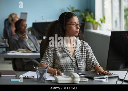 Young adult woman wearing headset working at computer in modern office, sitting at desk with telephone and stationery, focused on screen, colleagues visible in background Stock Photo