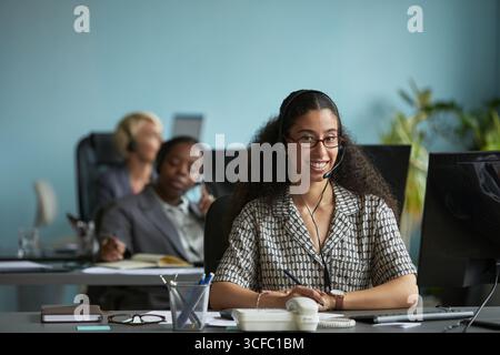 Portrait of young adult woman wearing headset smiling at desk in modern office, sitting in front of computer monitor with colleagues working in background, demonstrating customer support role Stock Photo