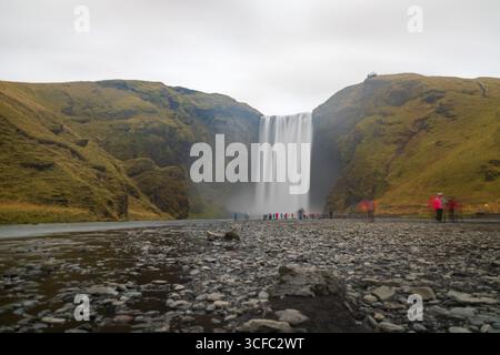 View of majestic Skógafoss waterfall cascading down rugged cliffs, with misty spray and blurred figures adding movement, captured on a cloudy day, Vik, Iceland. Stock Photo