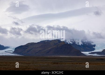 View of a vast, barren landscape with distant, rugged mountains shrouded in mist and ice, reflecting a serene yet powerful natural scene, Vik, Iceland. Stock Photo
