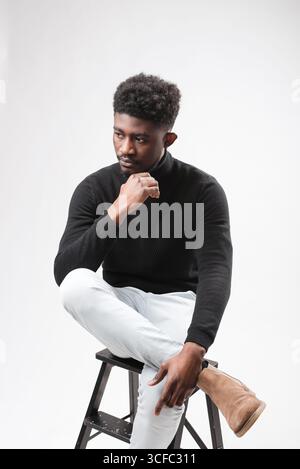 Relaxed curly bearded dark-skinned guy in casual outfit sitting at armchair, looking down over white studio background Stock Photo