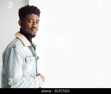 Serious young bearded guy in jeans jacket looking at camera while standing against white background with folded arms Stock Photo