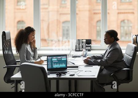 Two women, one Black and one biracial, working at office desk with laptops and documents, biracial woman talking on phone while Black woman reviewing paperwork Stock Photo