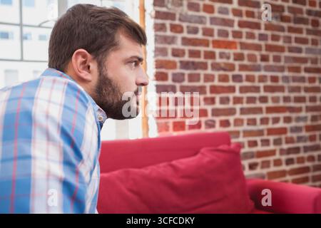 Man sitting on red sofa beside window in lounge with brick wall, gazing right, copy space Stock Photo