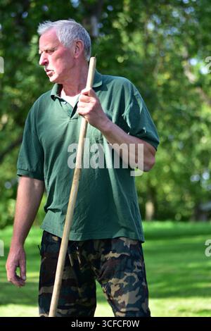 Close-up portrait of an elderly man in a green shirt and camouflage pants holding a stick outdoors, looking thoughtful in a bright countryside yard wi Stock Photo