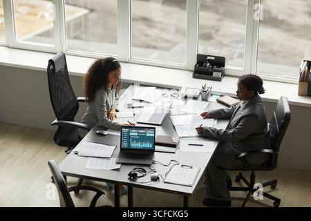 Two businesswomen, one Black and one biracial, working at office table, analyzing financial data on laptop and reviewing documents Stock Photo