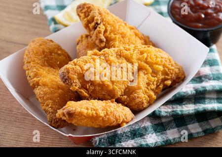 Close-up of golden crispy fried chicken tenders served in a red and white paper tray on wooden table. Stock Photo