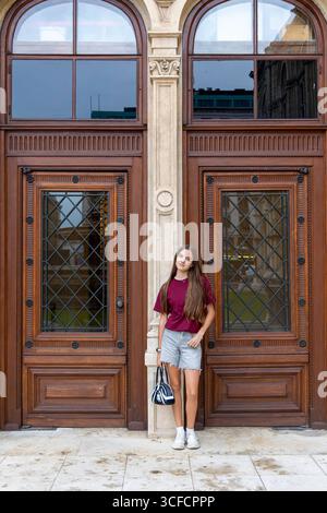 Young woman standing by ornate vintage doors. Portrait of a casually dressed young woman leaning against elegant wooden double doors with arched Stock Photo