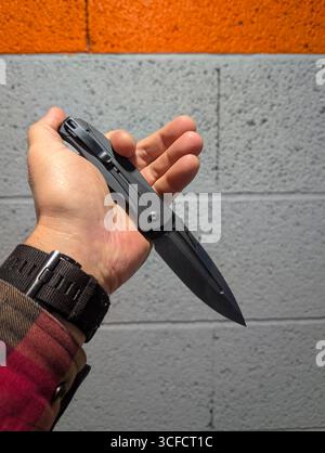A man's hand in a casual plaid shirt firmly holds an open black tactical folding knife, an essential everyday carry item. Stock Photo