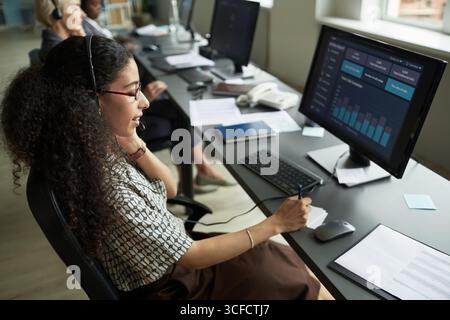 Young adult woman wearing headset working at computer desk analyzing data on monitor in modern office, colleagues sitting in background Stock Photo