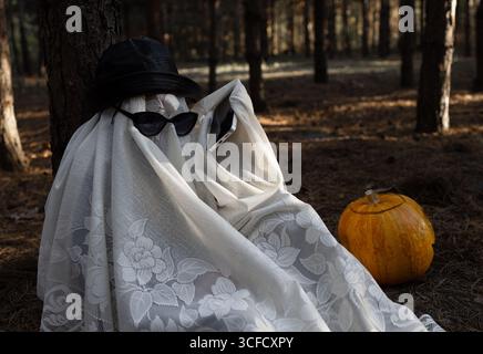A child in a white blanket and black glasses sits under a tree in a gloomy forest and talks on the phone. Children's creativity. Have fun on Halloween Stock Photo
