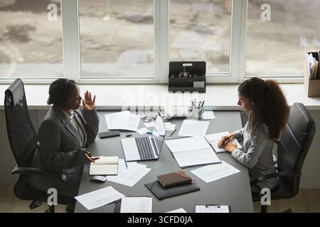 Two businesswomen, one Black and one biracial, sitting at office desk discussing documents and using laptop, both wearing headsets and engaging in professional conversation Stock Photo