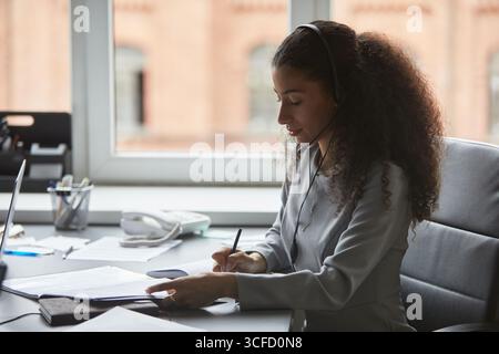 Young adult woman sitting at desk wearing headset, writing on documents while working in office environment, focused on paperwork with telephone and window in background Stock Photo