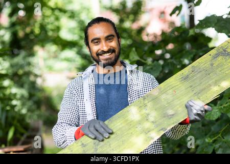 Smiling man carrying a wooden plank in a lush green garden. Germany Stock Photo