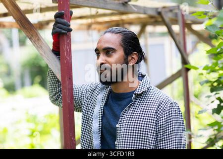 Man in checkered shirt and gloves stands thoughtfully outdoors by wooden structure. Germany Stock Photo